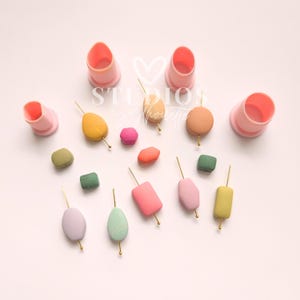 May include: A collection of colorful, geometric clay shapes on gold skewers, alongside pink, cylindrical cutters. The shapes include ovals, rectangles, and cubes in shades of pink, green, yellow, and orange. The word "STUDIOS" is visible in the background.