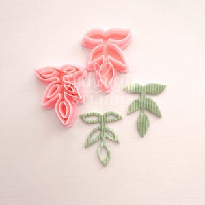 May include: Four leaf-shaped polymer clay cutters in shades of pink and green. The pink cutters have a smooth finish, while the green ones have a textured, striped appearance. The cutters are arranged on a light pink background.