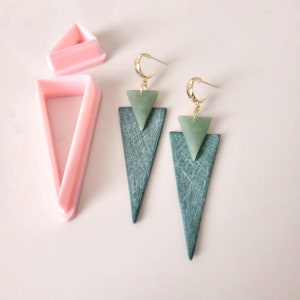 May include: A pair of green and gold triangle earrings. The earrings are made of two triangles, one on top of the other. The top triangle is a lighter shade of green and the bottom triangle is a darker shade of green. The earrings are suspended from gold hoops.