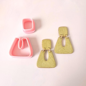 May include: Two pairs of gold-coloured earrings with geometric shapes. The earrings are made of clay and have a textured surface. The earrings are hanging from a pink surface.