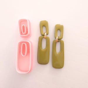May include: A pair of olive green statement earrings with a gold accent. The earrings are made of two rectangular pieces with a hole in the center. The earrings are shown next to a pink plastic mold.
