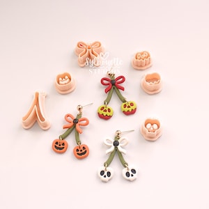 May include: Three pairs of dangle earrings shaped like cherries with jack-o'-lantern, skull, and apple faces, featuring bows and green stems. Accompanied by several peach-coloured plastic cookie cutters in various shapes, including a bow and pumpkin.