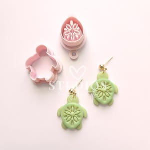 Könnte beinhalten: Ein Set mit zwei grünen Schildkrötenohrringen mit Blumenmuster und einem rosafarbenen Plastik-Schildkröten-Ausstecher.