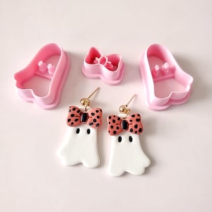 May include: A pair of white ghost-shaped earrings with orange and black polka dot bows. The earrings are made of polymer clay and have gold ear wires. The image also shows two pink plastic cookie cutters, one shaped like a ghost and the other shaped like a bow.