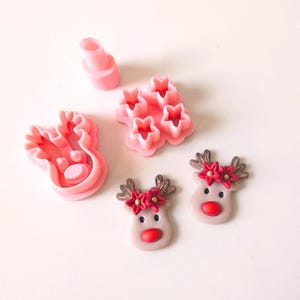 Puede incluir: Un juego de cortadores de galletas de plástico rosa para hacer formas de reno y estrella. El juego incluye dos cortadores de reno, un cortador de estrella y un pequeño cortador redondo. Los cortadores de reno tienen cuernos, una nariz y ojos. El cortador de estrella es una estrella de cinco puntas. El cortador redondo es pequeño y redondo.