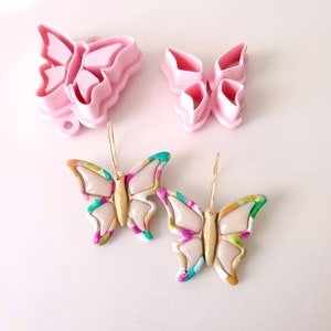 May include: Two pairs of butterfly earrings with gold hoops. The earrings are made of colorful polymer clay with a white background and gold accents. The earrings are handmade and unique.