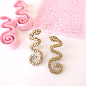 May include: Two gold-colored snake-shaped earrings with a textured surface. The earrings are made of a flexible material and have a wavy design.