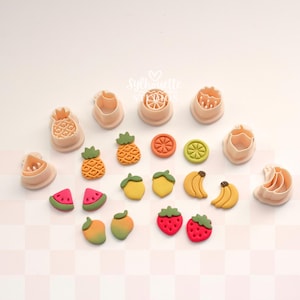 May include: A collection of peach-colored plastic fruit-shaped cookie cutters and their corresponding finished clay fruit pieces, including pineapple, watermelon, lemon, banana, mango, and strawberry shapes, arranged on a light checkered surface.