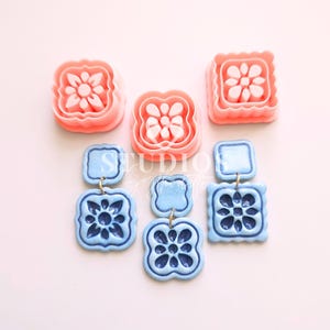 May include: Two pairs of blue and white ceramic earrings with a floral design. The earrings are made with two square shapes, one slightly larger than the other. The earrings are hanging from a gold hook.