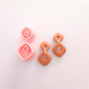 May include: Two pairs of earrings and two pink polymer clay cutters. The earrings are a warm peach and brown color with gold-colored metal accents. The cutters are pink and square-shaped with a rounded interior.