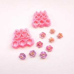 Könnte beinhalten: Rosa blütenförmige Ausstecher und mehrere kleine, mehrfarbige Blumendekorationen. Die Ausstecher gibt es in verschiedenen Größen und Formen, ideal zum Basteln. Die Blumen haben goldene Zentren und sind in Rosa- und Lilatönen gehalten.