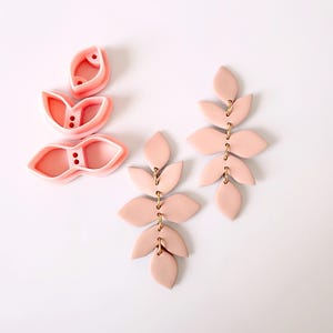 Puede incluir: Un par de pendientes de arcilla polimérica rosa en forma de hojas, conectados por anillos de salto dorados. Los pendientes se muestran junto a un juego de tres cortadores de hojas de arcilla polimérica rosa.