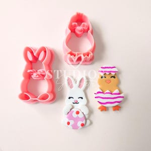 Pode incluir: Três cortadores de biscoitos de plástico rosa em forma de coelho da Páscoa, um pintinho em um ovo e um pintinho.