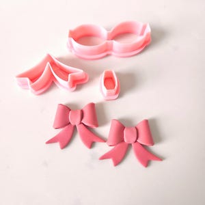 May include: Pink plastic cookie cutters in the shapes of a bow, a flower, and a pair of glasses. Two pink fondant bows are also shown.