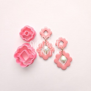 May include: Pink floral-shaped polymer clay cutters and matching earrings with pearl accents. The earrings have a gold-colored hook. The image is on a light pink background. The text "Sylvante Studios" is visible.