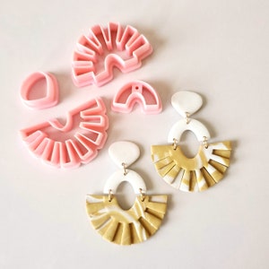 Könnte beinhalten: Zwei Paar handgefertigte Ohrringe aus Polymer Clay mit einem marmorierten Weiß- und Golddesign. Die Ohrringe haben ein fächerförmiges Design mit abgerundeter Oberseite und einer kleineren ovalen Form in der Mitte. Die Ohrringe sind aus rosa Polymer Clay-Ausstechern gefertigt.