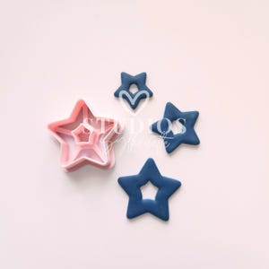 Puede incluir: Un conjunto de cortadores de galletas en forma de estrella y decoraciones en forma de estrella. Los cortadores de galletas son rosas y las decoraciones azul marino. La imagen está sobre un fondo rosa claro. El texto "STUDIOS by silhouette" es visible.