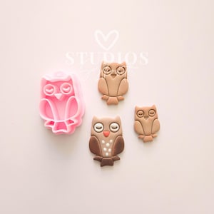 May include: A pink owl-shaped cookie cutter next to three brown owl-shaped cookies. The cookies have white and brown details. The cookie cutter is made of pink plastic. The cookies are in different sizes.