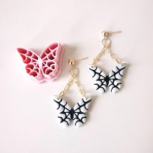 Puede incluir: Un par de pendientes colgantes con dijes de mariposa blanca con diseños de telaraña negra. Los pendientes están unidos a cadenas doradas y tienen postes de bola dorados.
