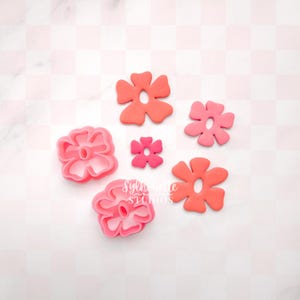 May include: Various flower-shaped cookie cutters in shades of pink and coral. Some cutters have a defined outline, while others are solid. The background features a pink and white checkered pattern. The image includes the text "Sylvie Studios".