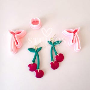 May include: A pair of cherry earrings with green stems and leaves. The earrings are made of polymer clay and have a gold-colored hook. The earrings are shown next to two pink plastic cookie cutters, one shaped like a heart and the other shaped like a bow.