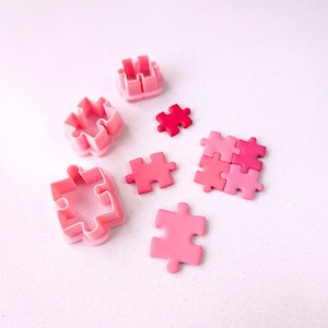 May include: A set of pink and red plastic puzzle-shaped cookie cutters and fondant pieces. The cutters are in different sizes and shapes. The fondant pieces are in various shades of pink.