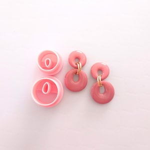 Puede incluir: Un par de pendientes de resina rosa con detalles dorados. Los pendientes tienen forma de dos círculos conectados. Los pendientes se muestran junto a dos moldes de plástico rosa.