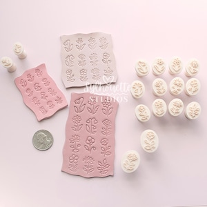 May include: A collection of light pink and off-white silicone stamps featuring various floral designs. The set includes three rectangular sheets with embossed flower patterns and several individual oval-shaped stamps, alongside a US quarter coin.