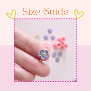 May include: A close-up of a finger holding a small purple flower with a yellow center. The flower is surrounded by other small flowers in various colors, including pink and purple.