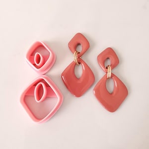 Könnte beinhalten: Ein Paar Statement-Ohrringe in Pink und Terrakotta mit geometrischem Design. Die Ohrringe sind aus Kunststoff und haben einen goldfarbenen Metallakzent.