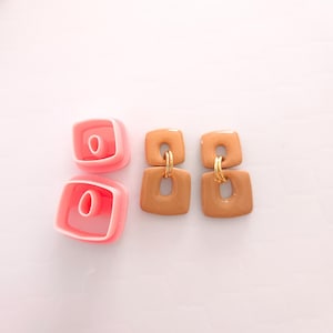 May include: Two pairs of brown square earrings with gold accents. The earrings are shown next to two pink plastic molds.