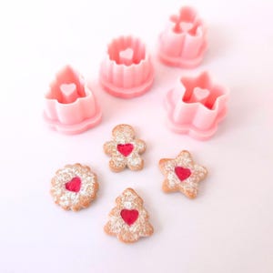 Puede incluir: Cinco cortadores de galletas de color rosa en forma de hombre de jengibre, estrella, árbol, corazón y círculo. Cuatro galletas con corazones de jalea roja están dispuestas sobre una superficie blanca. Las galletas tienen forma de hombre de jengibre, estrella, árbol y círculo.