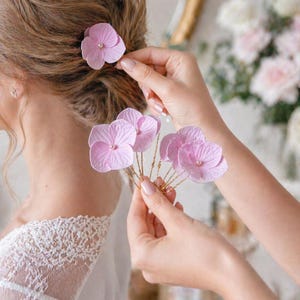 Può includere: I capelli di una persona sono adornati da delicate forcine a forma di fiori di ortensia rosa chiaro. Le forcine vengono inserite nei capelli. I fiori hanno un centro di perla e sono attaccati a steli di filo dorato. La persona indossa un abito di pizzo bianco.
