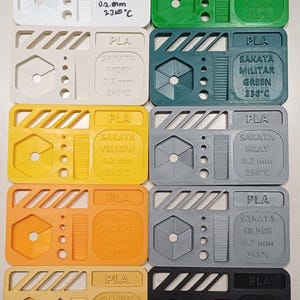 Puede incluir: Una colección de pruebas de impresión de filamento PLA multicolor. Cada muestra rectangular presenta un diseño único con texto que indica el tipo de filamento, el color y las especificaciones de impresión. Los colores incluyen blanco, verde, marfil, amarillo, naranja, gris, plata, dorado y negro.