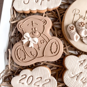 May include: A gift box filled with decorated cookies. The cookies are shaped like a cloud, a teddy bear, and a circle. The cloud cookies have the words "Fufu" and "2022" on them. The circle cookie has the word "Baby" on it. The teddy bear cookie is brown and has a white bow.