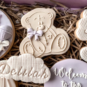 May include: A set of decorated cookies in a gift box. The cookies are decorated with a teddy bear, a cloud with the name "Delilah" on it, and a purple cookie that says "Welcome to the World".