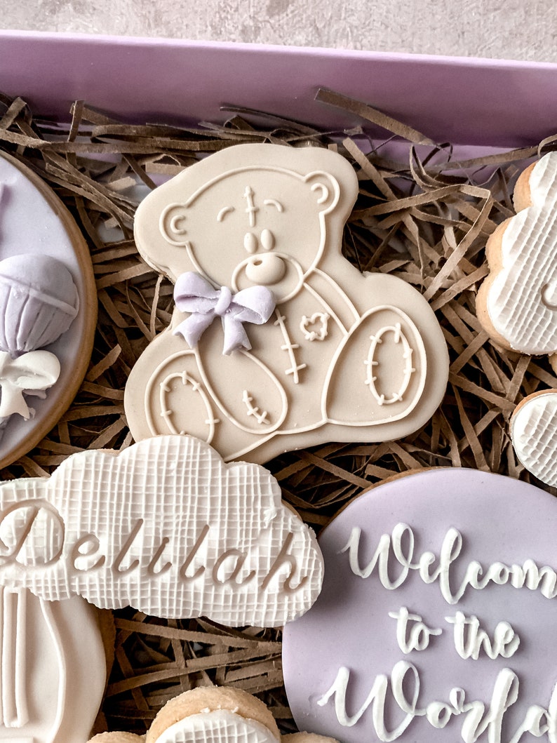 May include: A set of decorated cookies in a gift box. The cookies are decorated with a teddy bear, a cloud with the name "Delilah" on it, and a purple cookie that says "Welcome to the World".