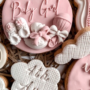 May include: A set of pink and white decorated cookies with the text "Baby Girl" and "4th Aug" on two of the cookies. The cookies are arranged on a brown paper background.