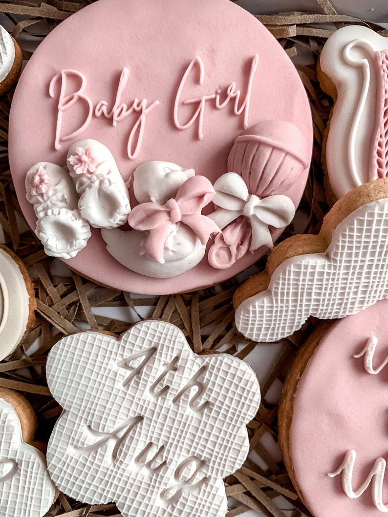 May include: A set of pink and white decorated cookies with the text "Baby Girl" and "4th Aug" on two of the cookies. The cookies are arranged on a brown paper background.