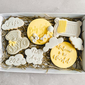 May include: A white gift box filled with yellow and grey decorated cookies. The cookies have various designs including a teddy bear, a cot, clouds, and the words "Baby Girl", "Welcome to the World", "4th Aug", and "2022".