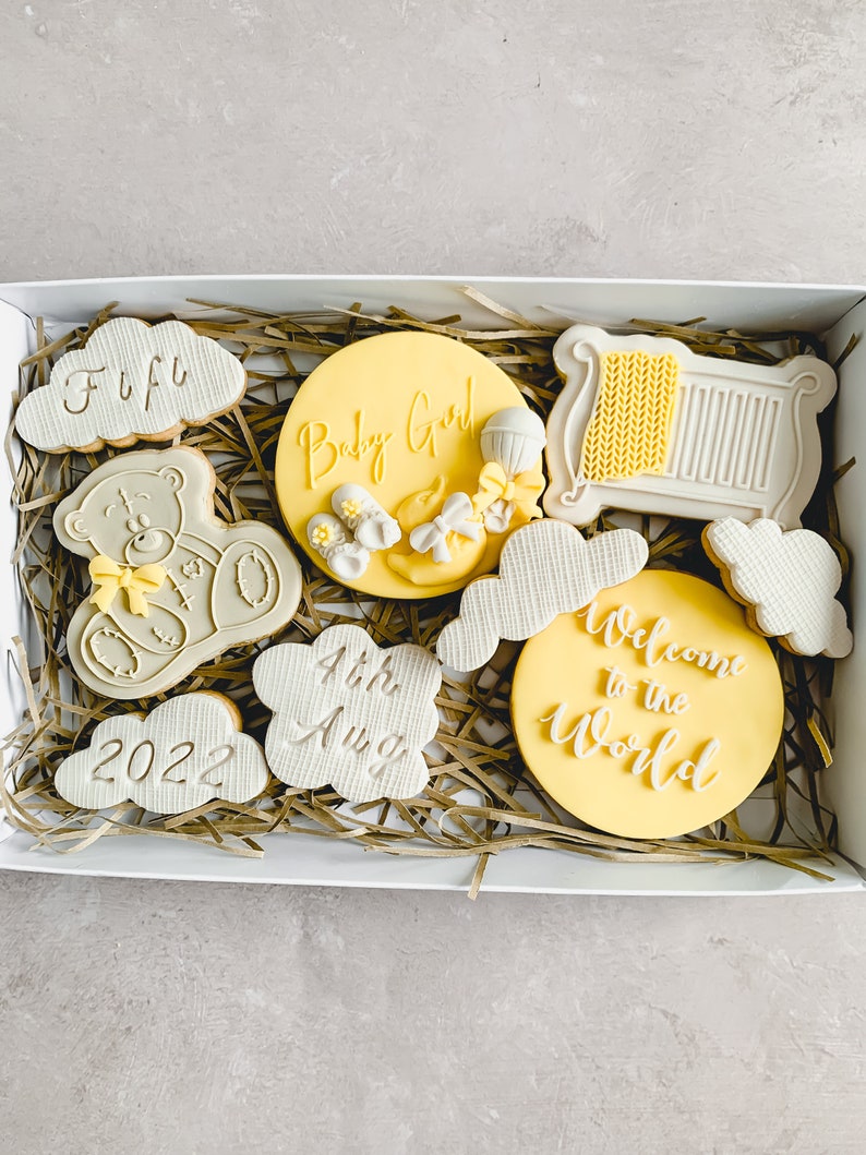 May include: A white gift box filled with yellow and grey decorated cookies. The cookies have various designs including a teddy bear, a cot, clouds, and the words "Baby Girl", "Welcome to the World", "4th Aug", and "2022".