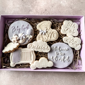 May include: A gift box filled with decorated sugar cookies. The cookies are decorated with purple and white icing and feature designs such as a teddy bear, a cot, clouds, and the text "Baby Girl", "Delilah", "Welcome to the World", and "8th Feb 2022".