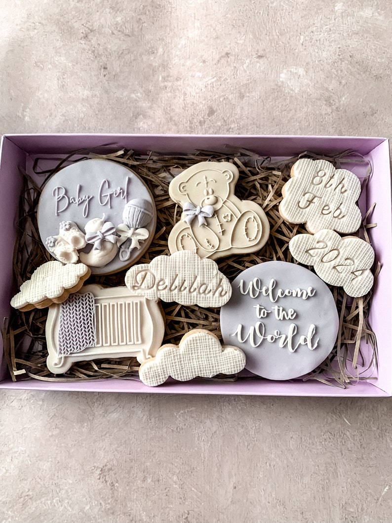 May include: A gift box filled with decorated sugar cookies. The cookies are decorated with purple and white icing and feature designs such as a teddy bear, a cot, clouds, and the text "Baby Girl", "Delilah", "Welcome to the World", and "8th Feb 2022".