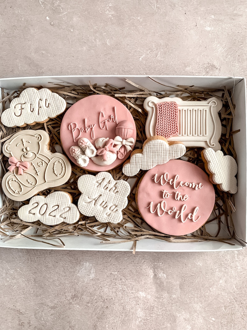 May include: A white gift box filled with pink and white decorated cookies. The cookies are shaped like clouds, a teddy bear, a crib, and a circle. The cookies have text on them, including "Baby Girl", "Welcome to the World", "4th Aug", and "2022".