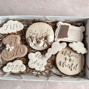 May include: A gift box filled with brown and white decorated cookies. The cookies are shaped like clouds, a teddy bear, a crib, and a circle. The cookies are decorated with the words "Baby Girl", "Welcome to the World", "4th Aug", "2022", and "Fufu".