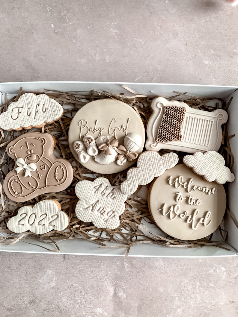 May include: A gift box filled with brown and white decorated cookies. The cookies are shaped like clouds, a teddy bear, a crib, and a circle. The cookies are decorated with the words "Baby Girl", "Welcome to the World", "4th Aug", "2022", and "Fufu".