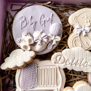 May include: A gift box filled with decorated cookies for a baby shower. The cookies are decorated with a teddy bear, a cot, clouds, and a cookie that says "Baby Girl" with a rattle and a baby shoe.