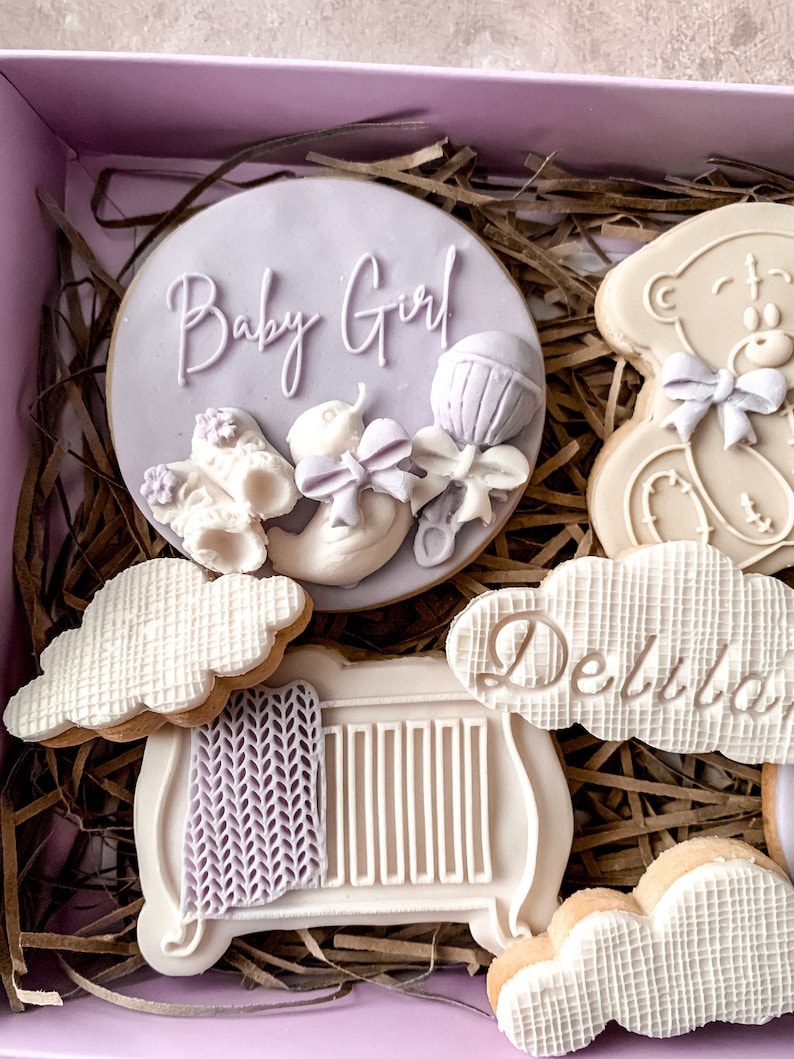May include: A gift box filled with decorated cookies for a baby shower. The cookies are decorated with a teddy bear, a cot, clouds, and a cookie that says "Baby Girl" with a rattle and a baby shoe.