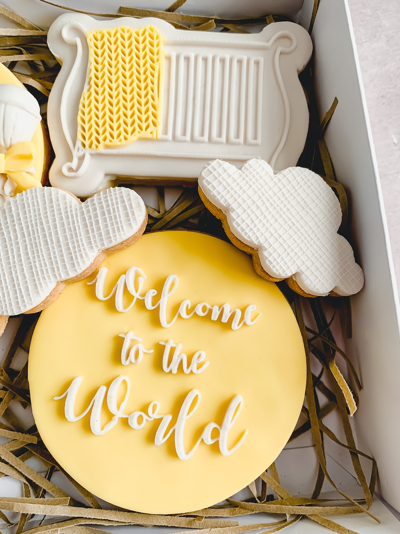 May include: A set of four yellow and white decorated cookies in a box. The cookies are shaped like a cot, a cloud, and a circle with the text "Welcome to the World".