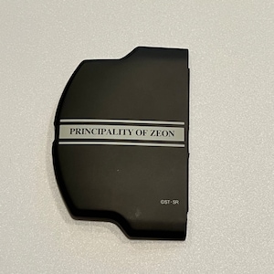May include: A black plastic case for a handheld video game console. The case has a silver stripe with the text "PRINCIPALITY OF ZEON" printed on it. The text "©ST-SR" is printed in the lower right corner.